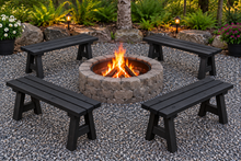 Load image into Gallery viewer, Four black outdoor bench chairs arranged around a round stone fire pit with visible flames, set on a gravel patio in a landscaped garden area with plants and small lights.