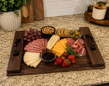 Load image into Gallery viewer, A dark wood charcuterie serving board with black handles, styled with sliced cheeses, cured meats, crackers, olives, grapes, strawberries, and a small bowl of preserves on a granite kitchen countertop.