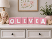 Load image into Gallery viewer, Personalized pink children's letter blocks spelling the name 'OLIVIA' with white letters, displayed on a white dresser in a styled nursery setting with a plush bunny, lamp, framed floral print, and eucalyptus plant.
