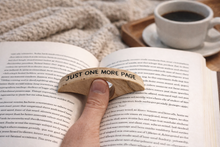 Load image into Gallery viewer, A hand holding a curved wooden bookmark or sign that reads 'JUST ONE MORE PAGE' over an open book with printed text, with a coffee cup in the blurred background.
