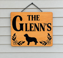 Load image into Gallery viewer, A personalized wooden hanging sign with the text 'THE GLENN'S' in black lettering, a black dog silhouette, and black floral scroll details, suspended by a black chain against white siding.
