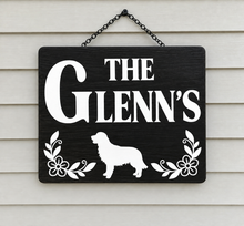 Load image into Gallery viewer, A personalized black wooden hanging sign with white text reading 'THE GLENN'S' and a white dog silhouette, flanked by white floral motifs, hanging against a light beige siding background.
