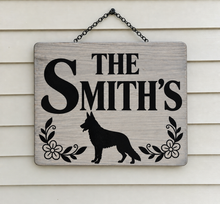 Load image into Gallery viewer, A personalized wooden hanging sign with a weathered gray finish, featuring the text 'THE SMITH'S' in black, a black dog silhouette, and black floral motifs. The sign hangs from a black metal chain against a white horizontal siding background.

