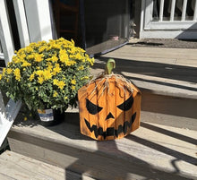 Load image into Gallery viewer, A rustic orange wooden jack-o'-lantern decoration with black painted facial features sits on a wooden porch next to a potted yellow flower.
