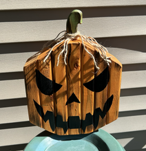 Load image into Gallery viewer, A rustic wooden jack-o'-lantern Halloween decoration with a carved face, hanging twine stem, and a painted green top, shown outdoors against siding.
