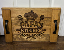 Load image into Gallery viewer, A rectangular wooden serving tray or decorative board with two black metal handles, featuring a dark engraved design that reads 'PAPA'S KITCHEN' with a chef's hat, utensils, and crossed tools. The tray is photographed standing upright against a white paneled wall on a dark wood floor.

