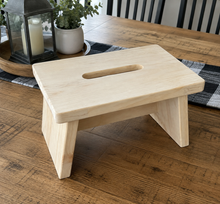Load image into Gallery viewer, A small light natural wood step stool with a central cutout, shown on a wooden table in a home setting with a lantern, plant, and black-and-white buffalo plaid runner in the background.
