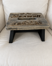 Load image into Gallery viewer, A rustic wooden step stool with a weathered gray-brown top featuring engraved racing car and checkered flag designs, supported by four black legs, shown on a beige upholstered chair.

