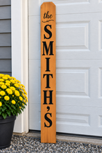 Load image into Gallery viewer, A personalized wooden sign reading 'the SMITH'S' in black lettering, leaning against a white garage door next to a potted plant with yellow flowers.
