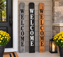 Load image into Gallery viewer, Three vertical wooden 'WELCOME' sign boards leaning against a beige house exterior door, flanked by yellow flower pots and a lit lantern.
