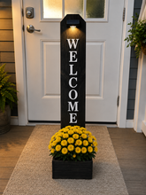 Load image into Gallery viewer, A tall black wooden welcome sign with white lettering stands on a front porch beside a white door, with a rectangular planter holding bright yellow flowers at the base.
